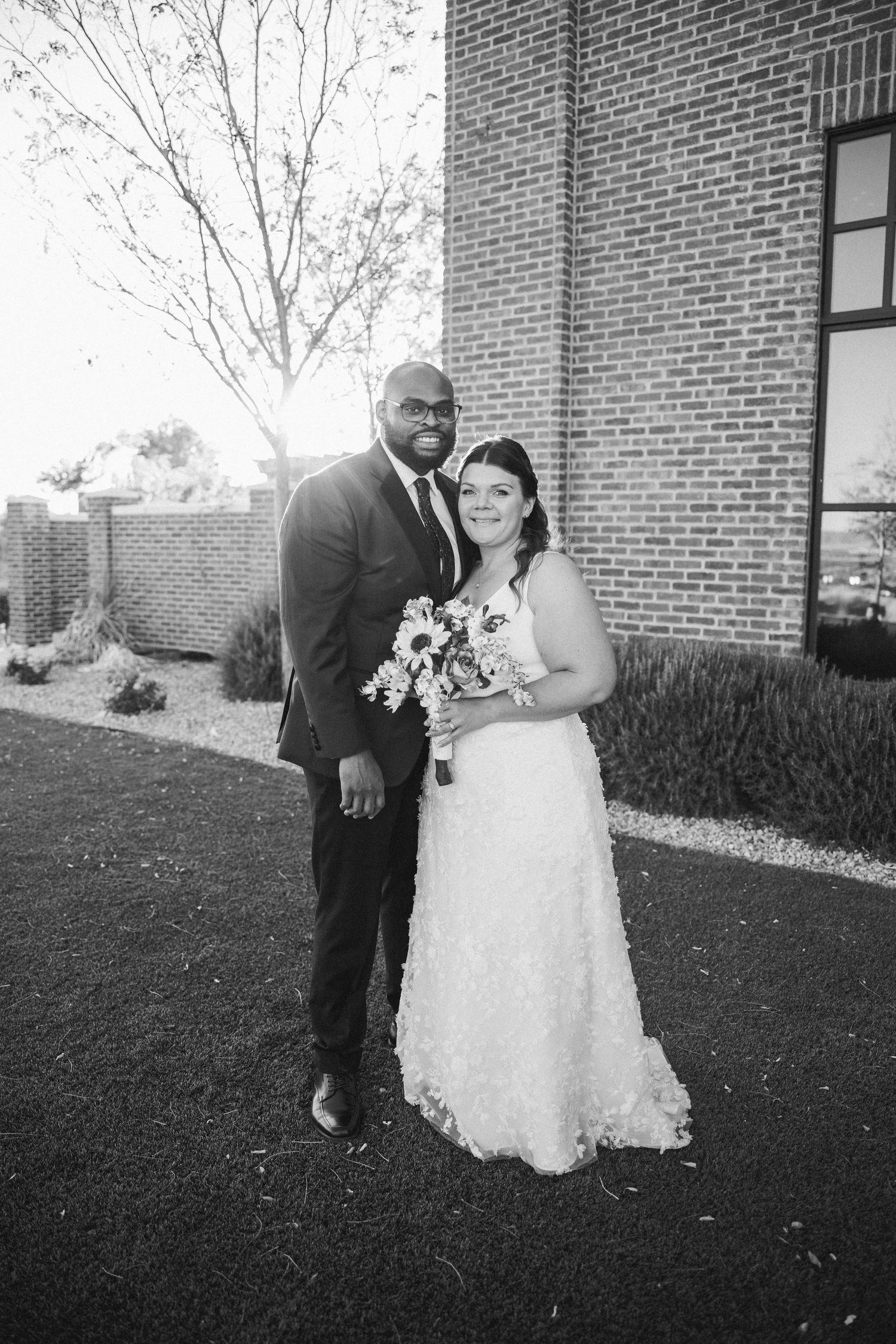 A bride and groom pose together outside a brick building on their wedding day.