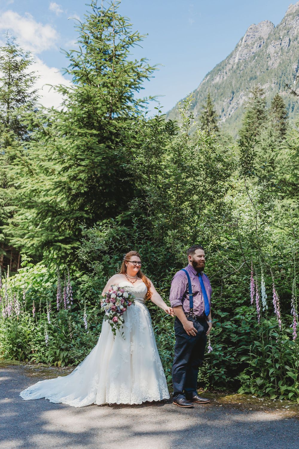 Bride stands behind groom at their first look surrounded by flowers and mountains