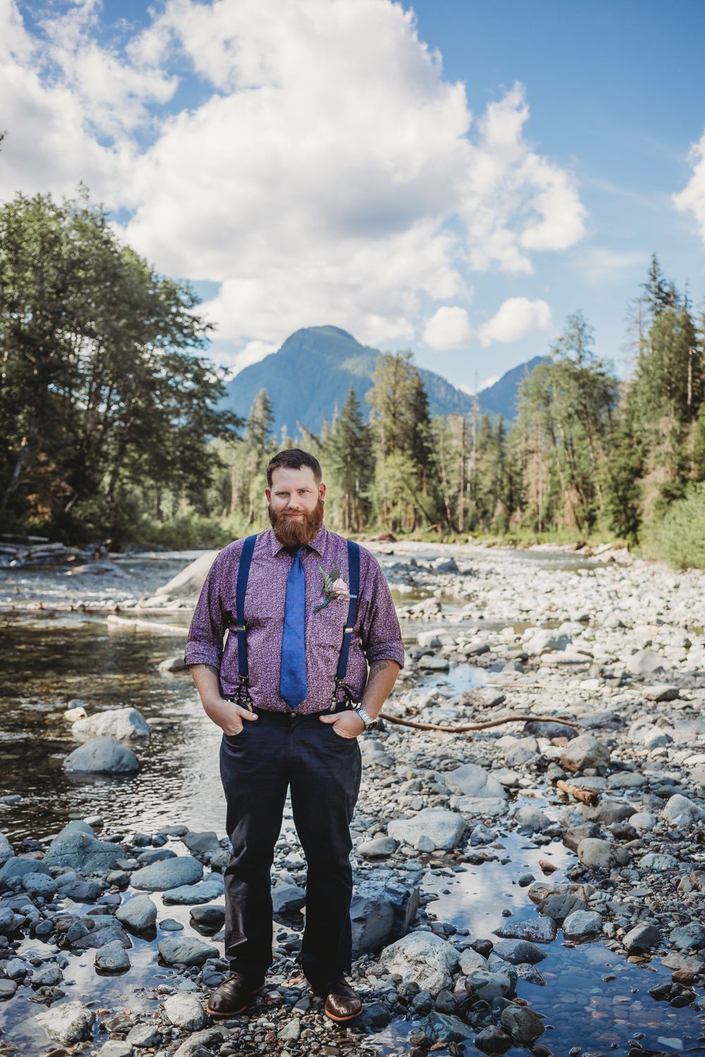 Groom stands with hands in his pocket along the river in the mountains at his elopement