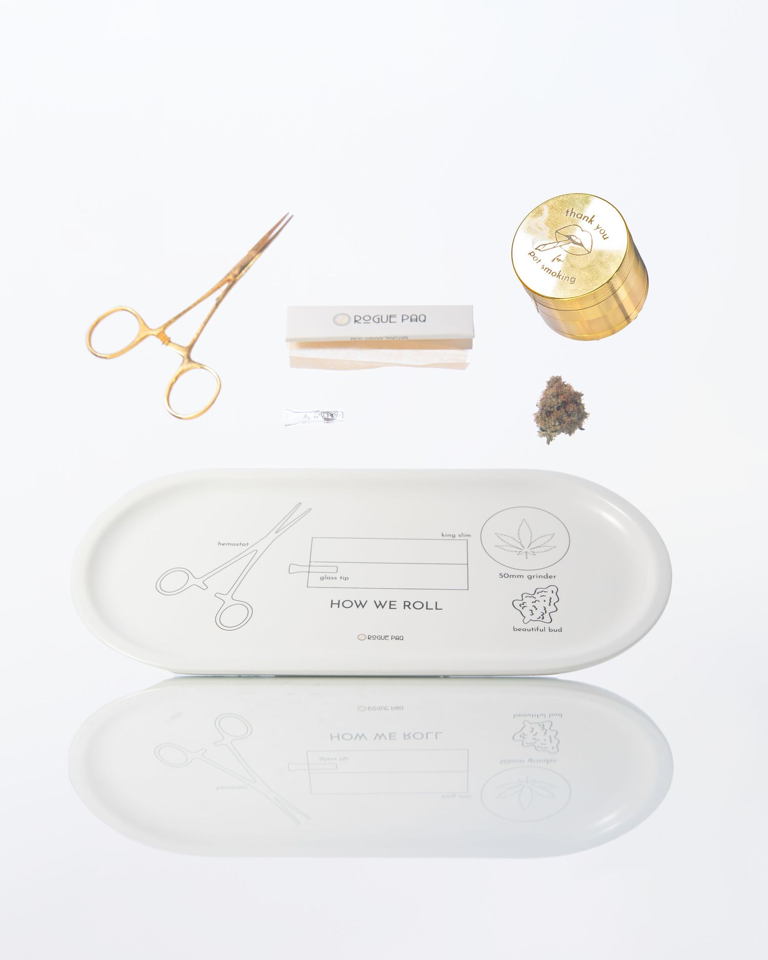 Flat lay of cannabis rolling accessories including grinder, papers, roach clip on white tray.