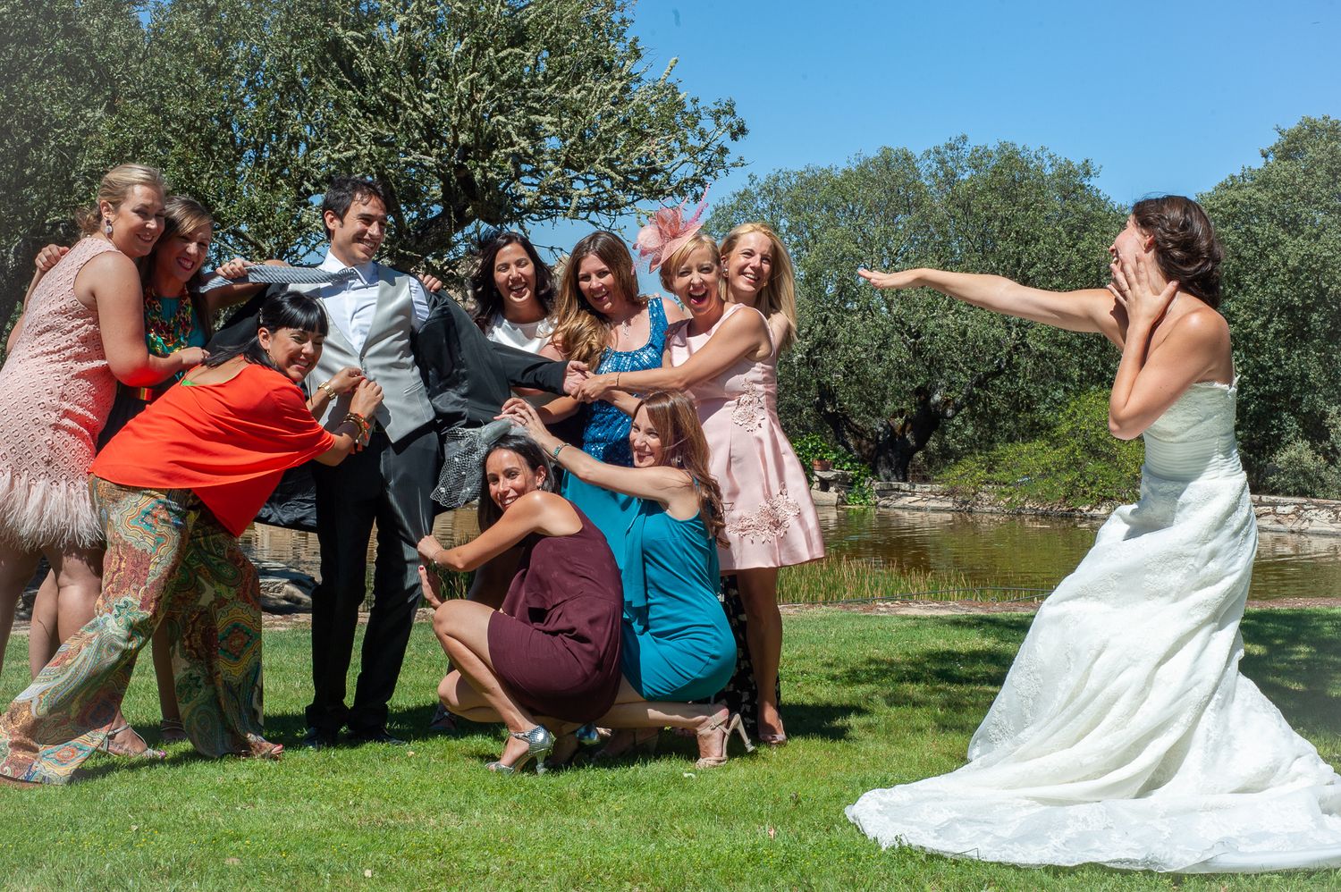 Bride playfully tosses bouquet to excited wedding guests gathered outdoors near pond on sunny day.