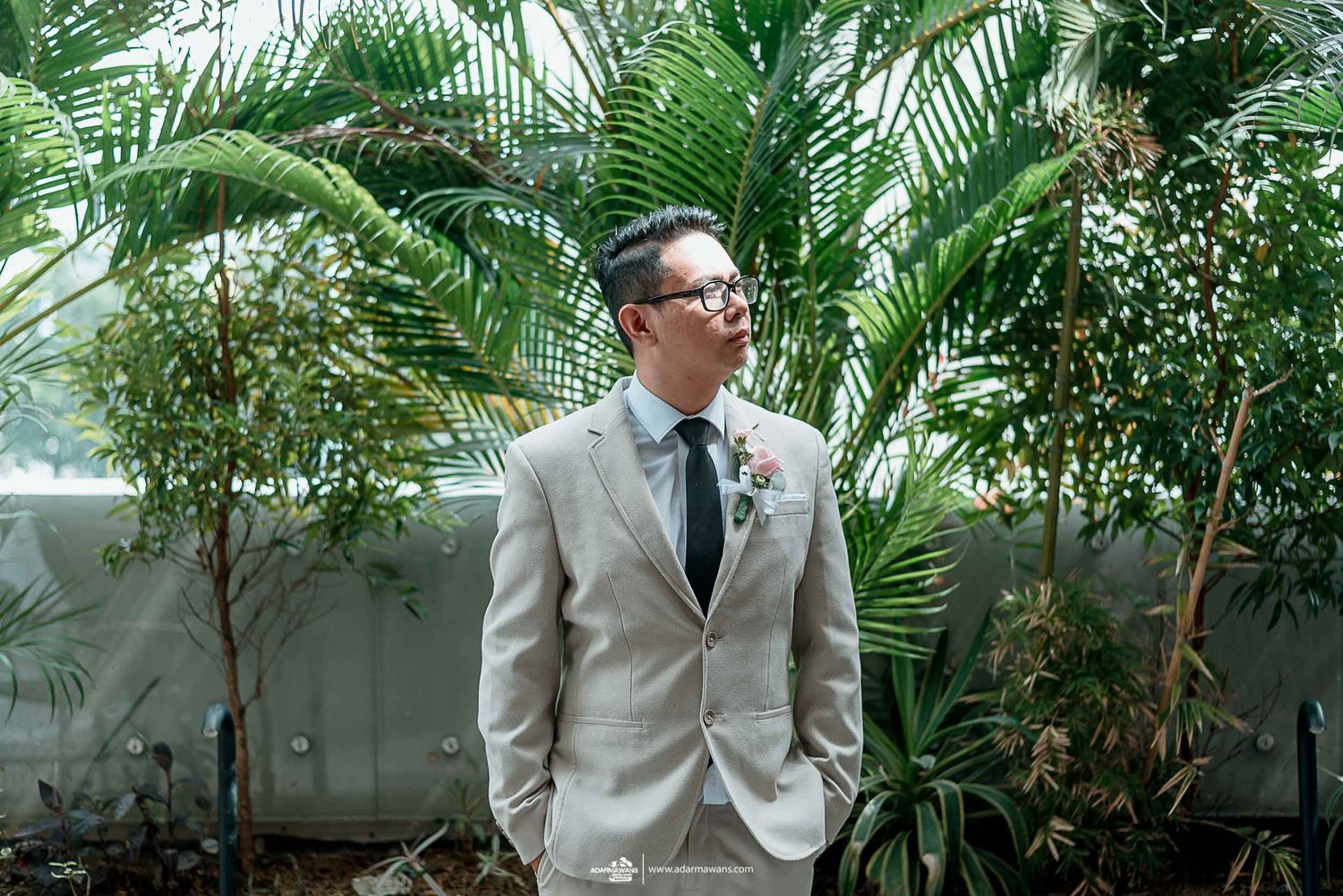 Groom in light gray suit and glasses poses in front of tropical palm plants.