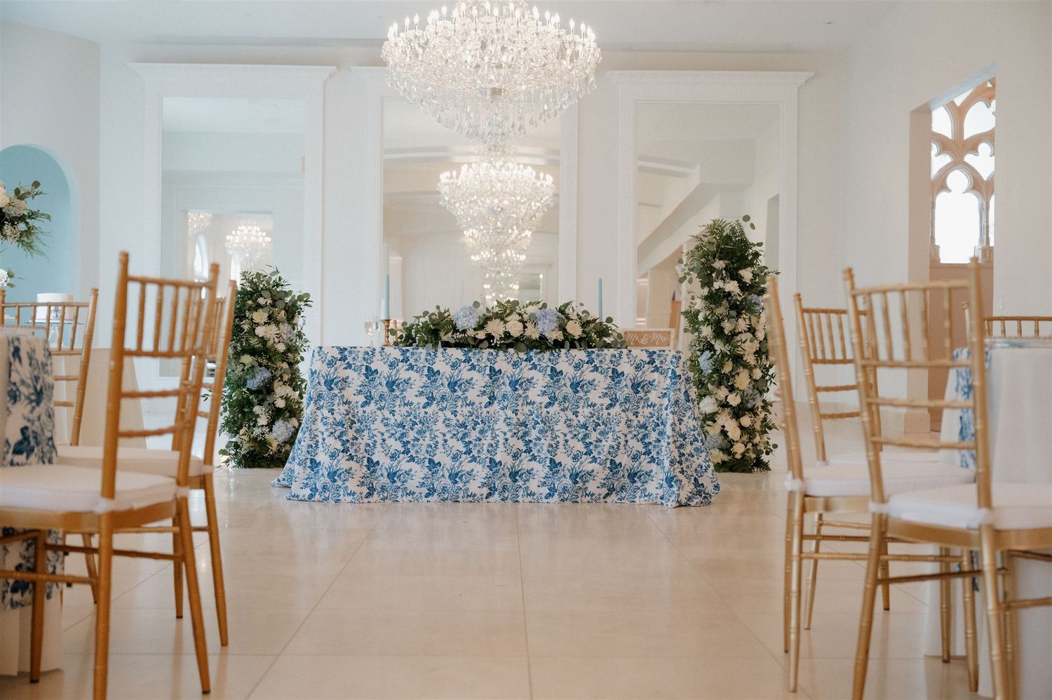 Wedding reception setup with blue floral tablecloth, crystal chandeliers and gold chairs.