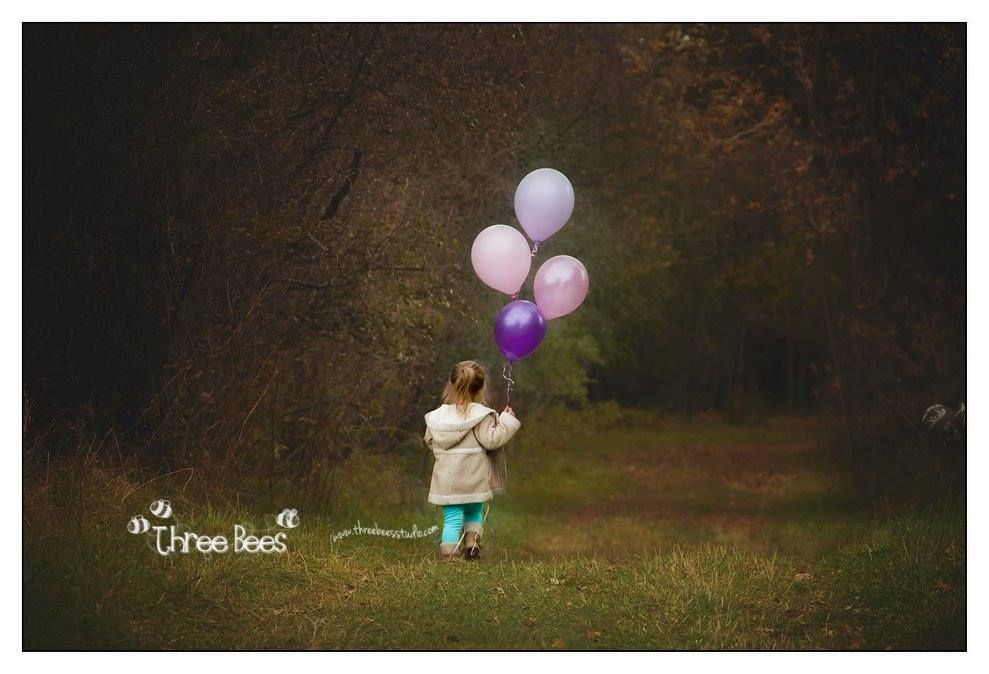 A young child in a white dress holding purple and pink balloons in a dark moody forest setting.