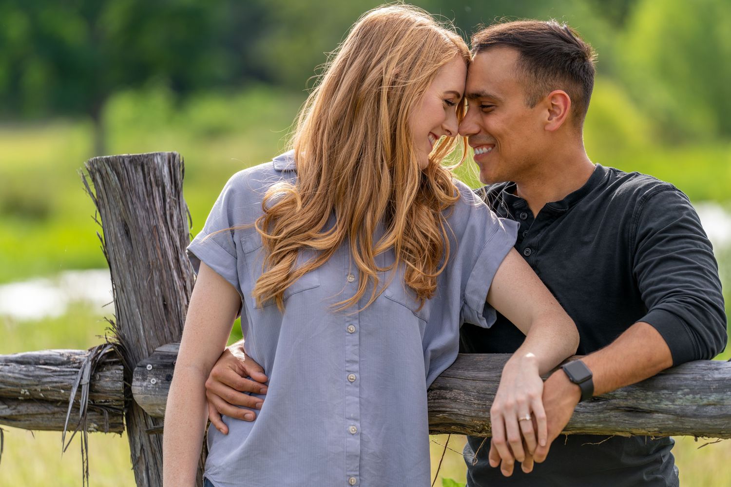 Intimate moment between couple leaning on wooden fence in sunny countryside setting.