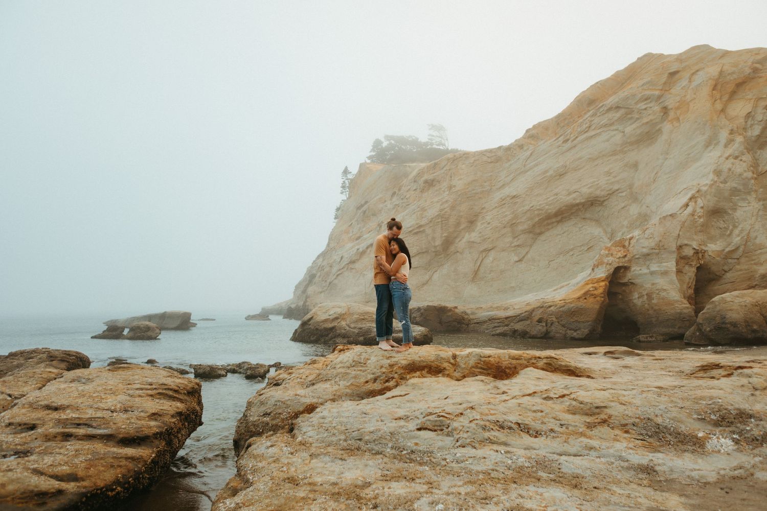 Person stands on rocky coastal cliffs overlooking misty ocean landscape.