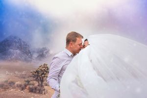 A romantic wedding photo of a couple embracing in a desert landscape with mountains and dreamy purple sky.