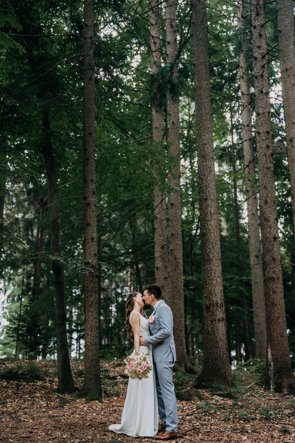 Romantic wedding couple embraces in a lush forest setting surrounded by tall evergreen trees.