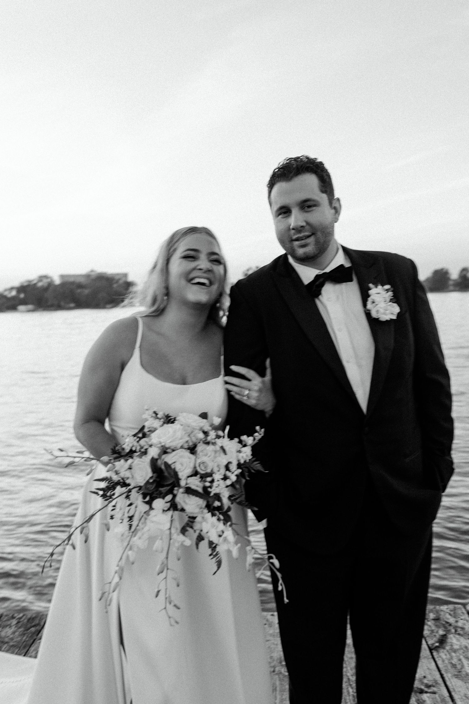 Joyful black and white wedding portrait by the water at sunset with elegant bouquet.