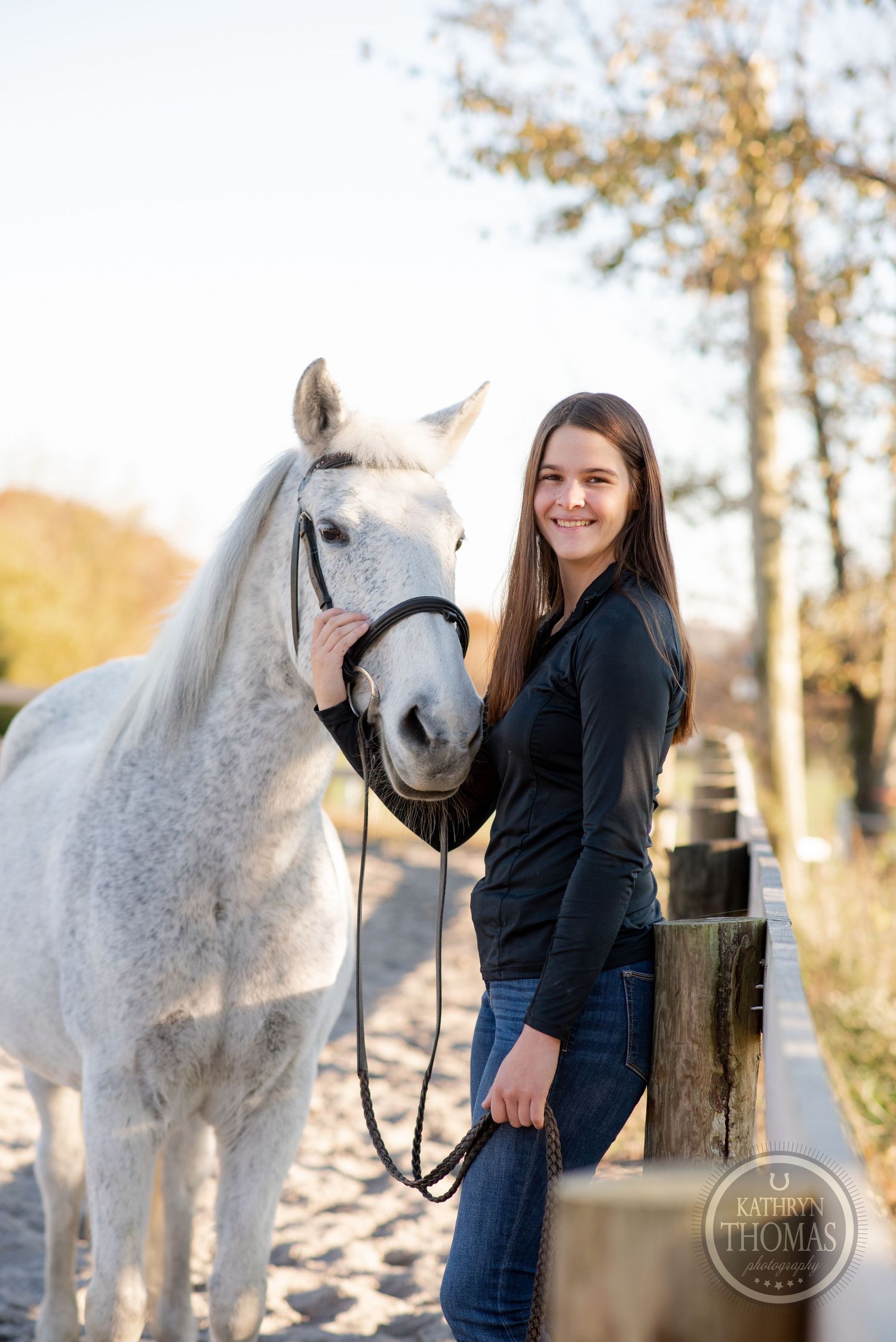 A majestic white horse poses with their handler by a wooden fence on a sunny autumn day.