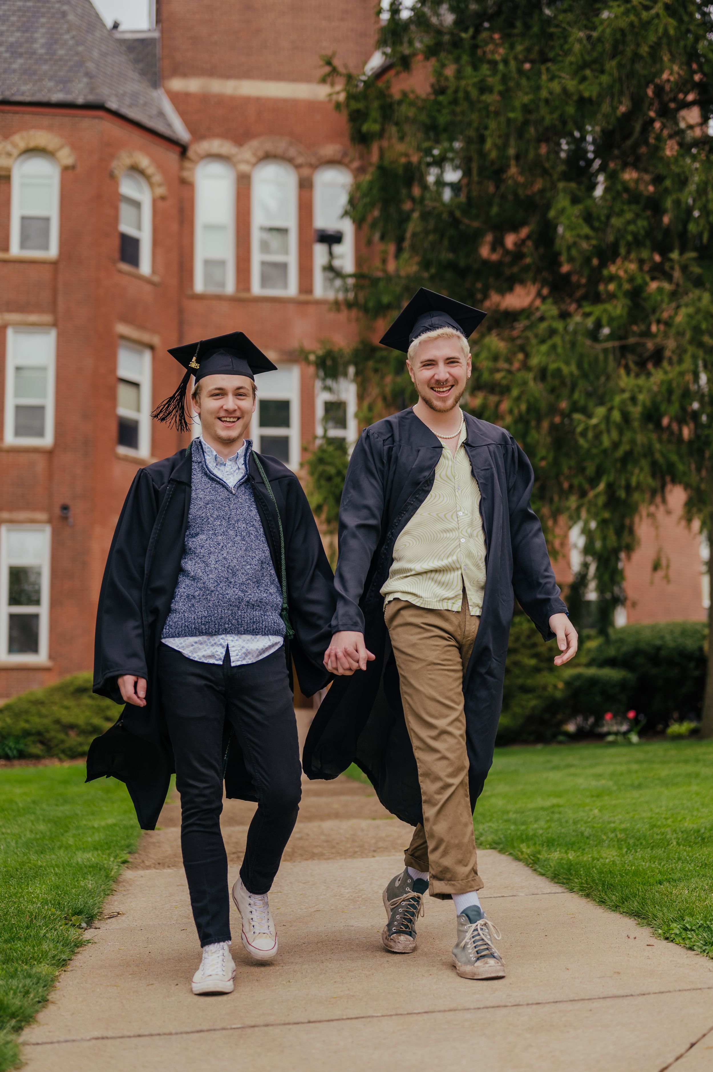 Two proud Slippery Rock University graduates, holding hands in unity, walk toward the camera on campus, radiating joy.