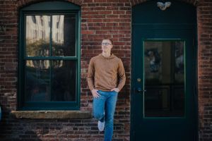 A male senior leans against a downtown Akron brick wall, gazing into the distance with an air of cool composure.