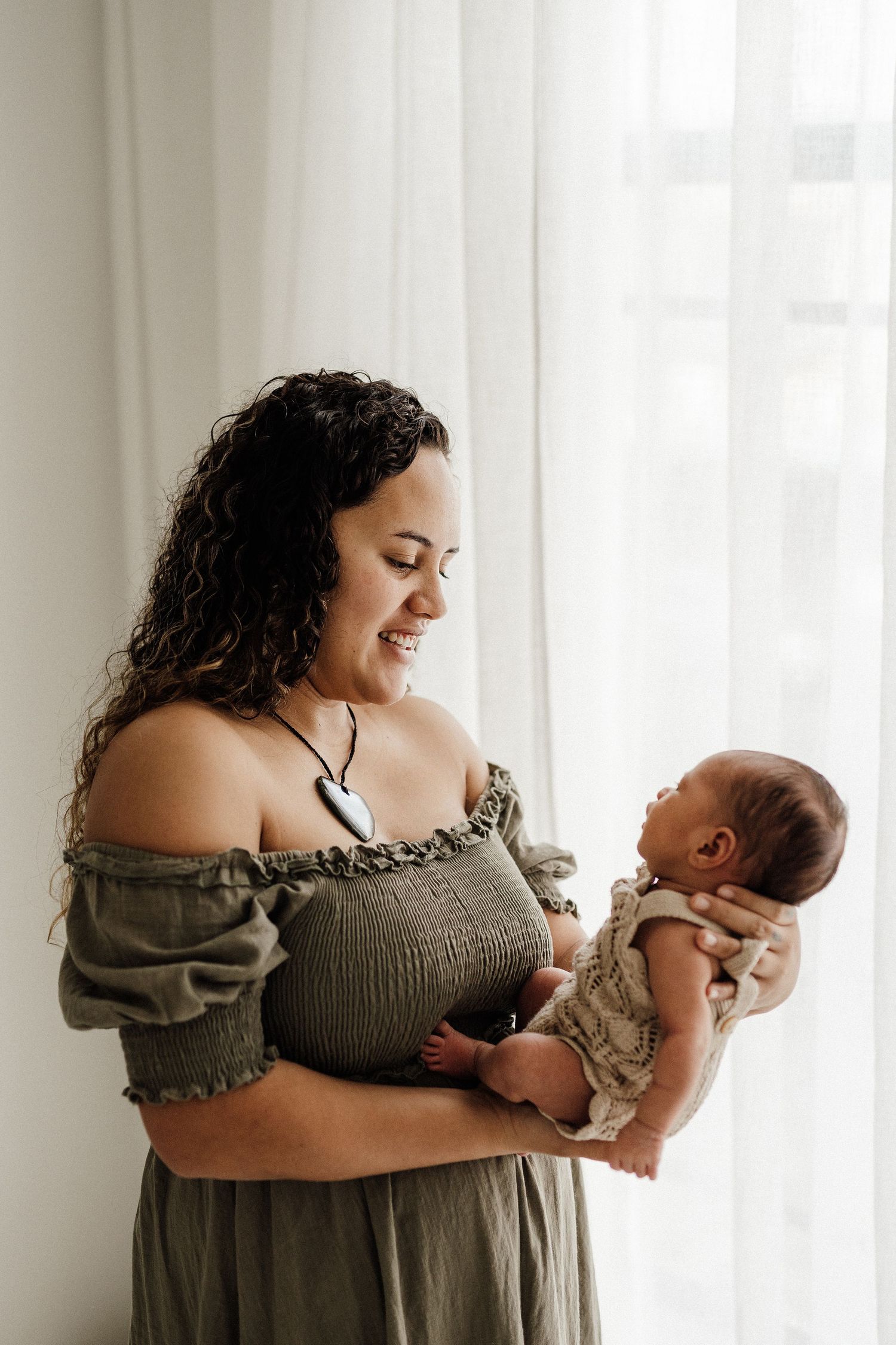 Mother in olive green dress holds and smiles at newborn baby by window in natural light portrait.