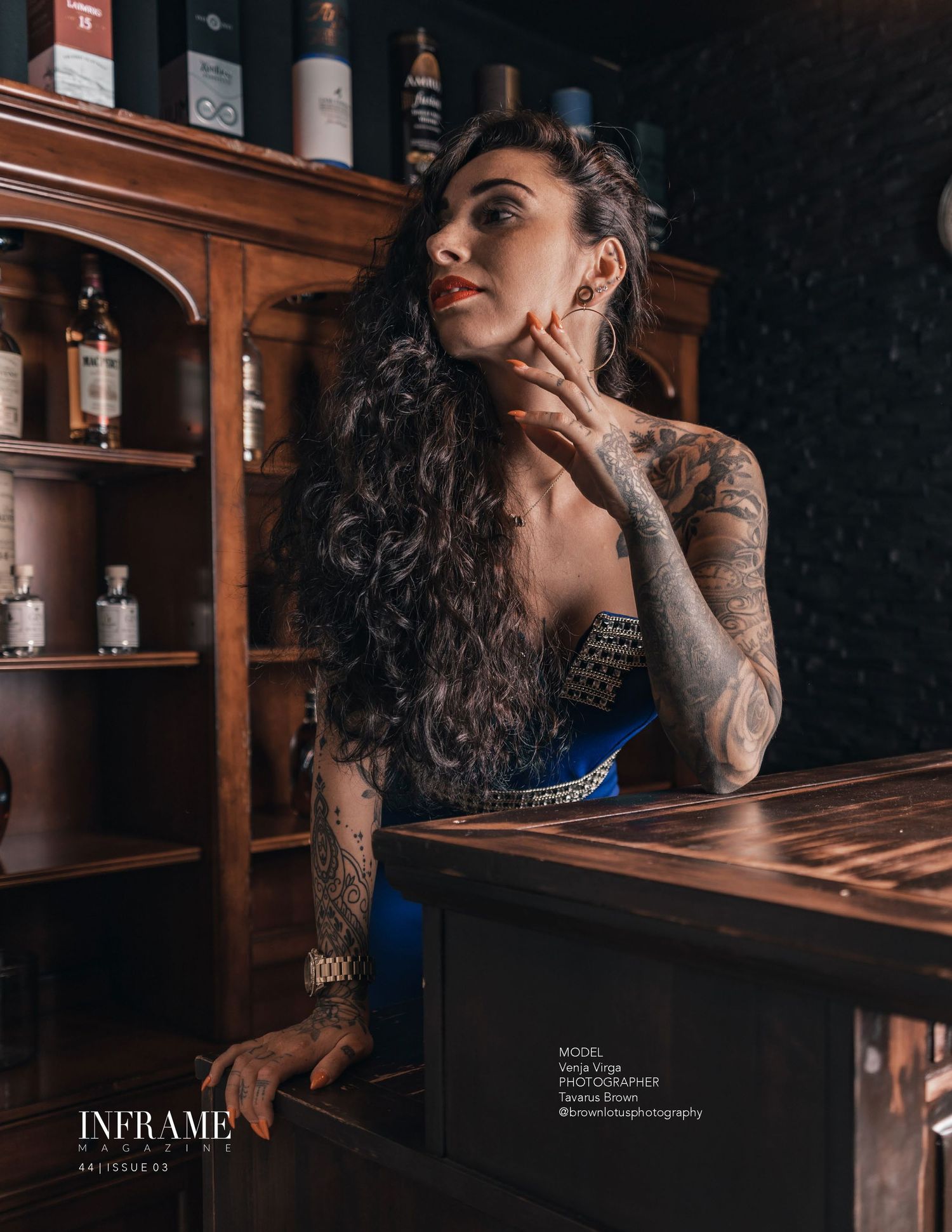 Tattooed woman in blue corset leaning on wooden bar counter with liquor bottles.