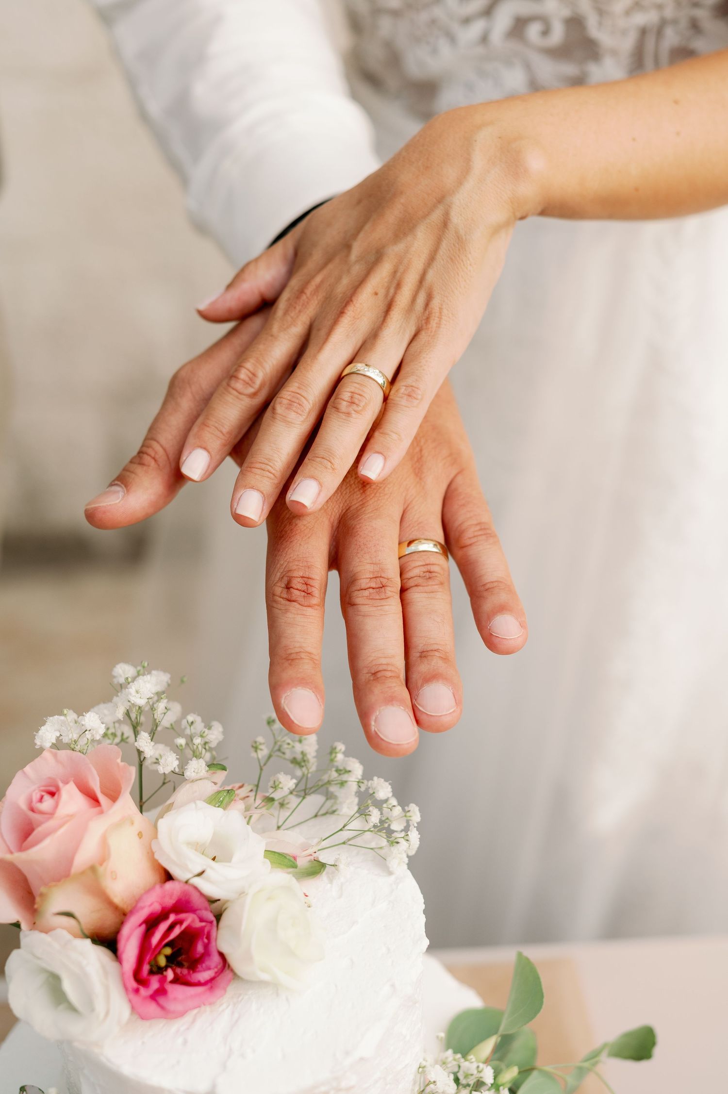 Close-up of wedding rings and hands with pink and white floral cake decoration.