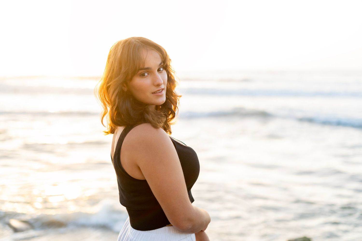 A high school senior in black top gazing at sunset over ocean waves during golden hour.
