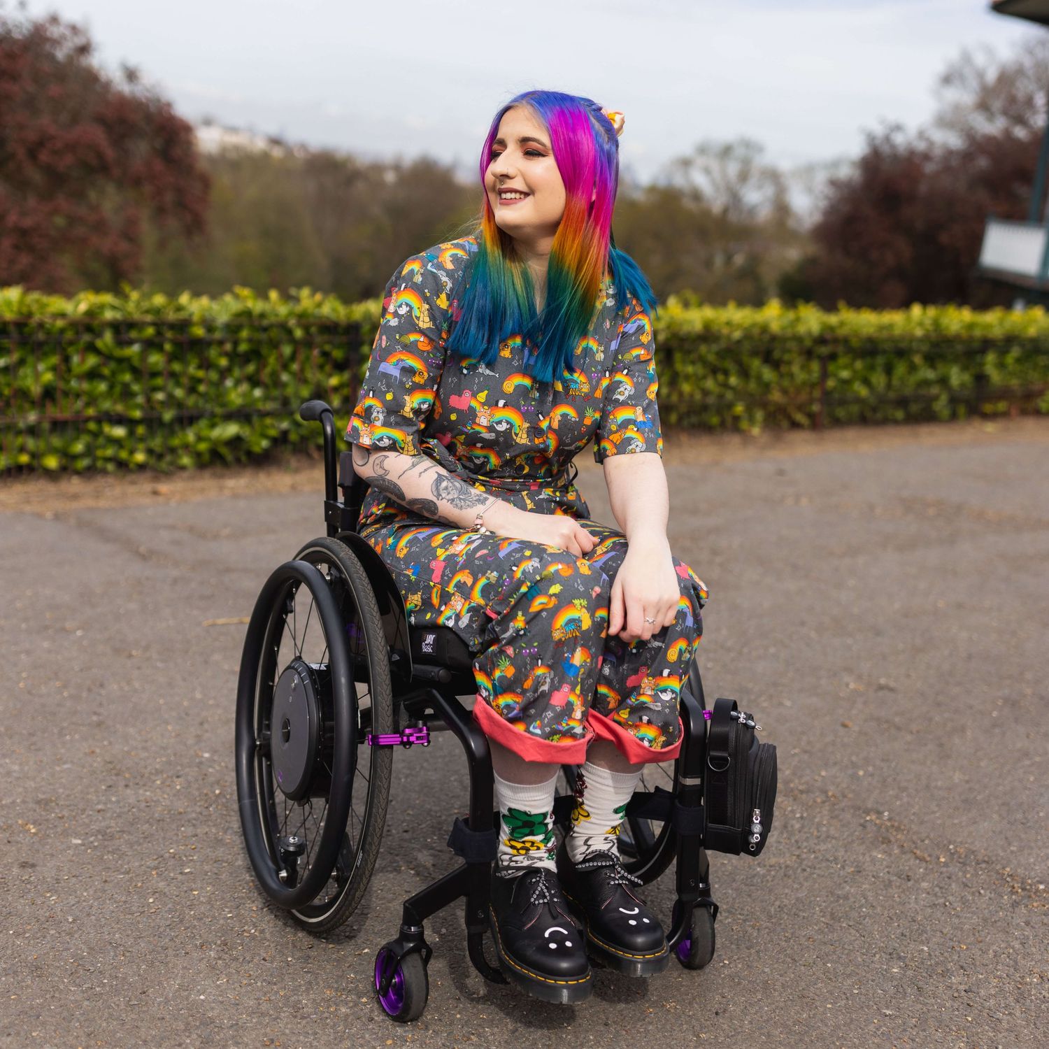 London rainbow haired disabled influencer posing sat smiling in their wheelchair wearing a rainbow outfit.