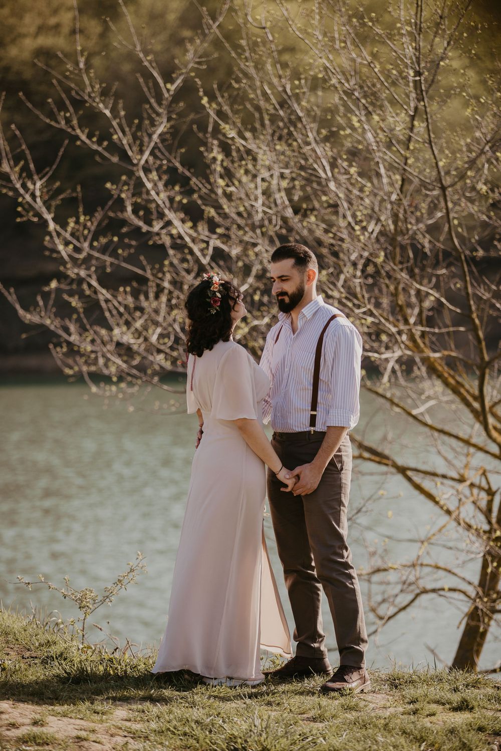 A couple stands by a lake surrounded by bare branches in romantic evening light.
