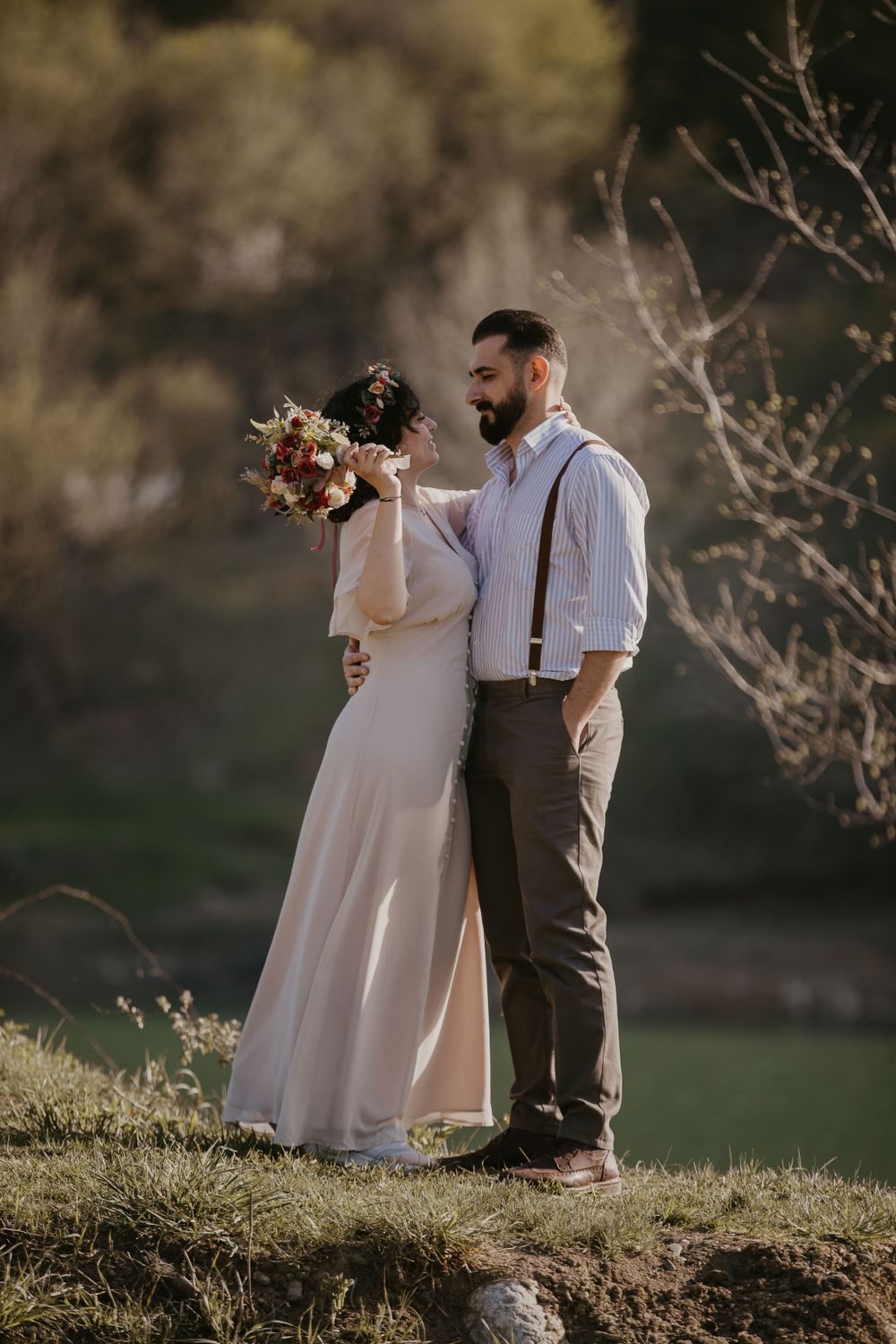 A couple in elegant attire embraces on a hillside with a bouquet of flowers.