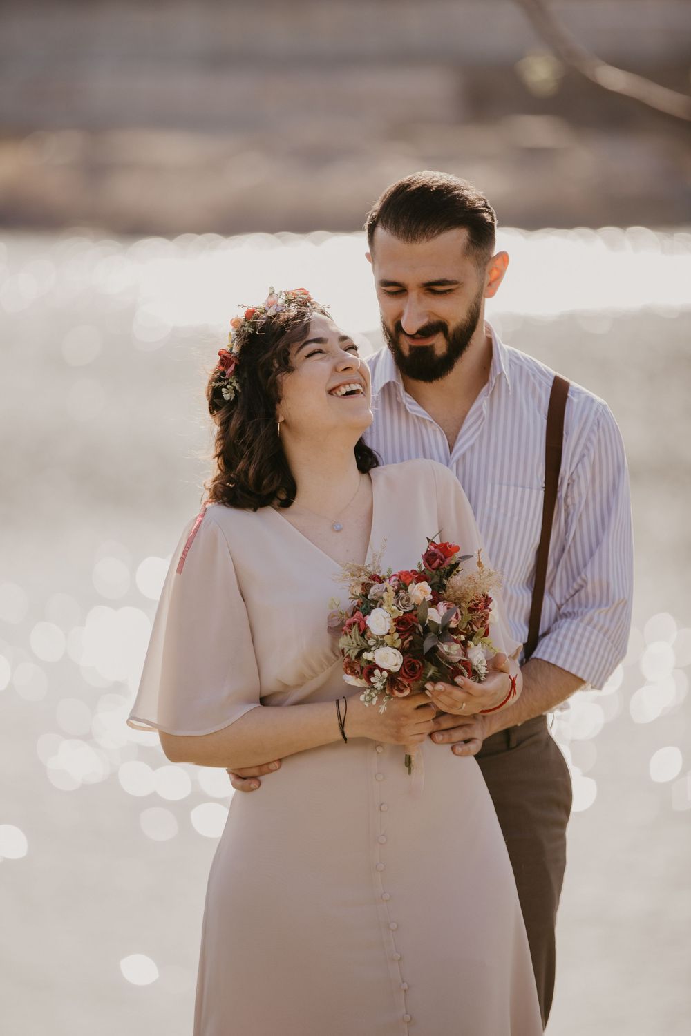 A couple laughs together while holding a bouquet with sparkling water in the background.