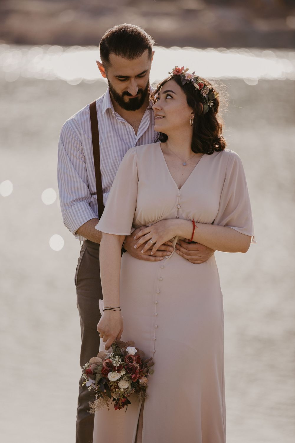 A couple shares an intimate moment while holding a floral bouquet against sparkling water.