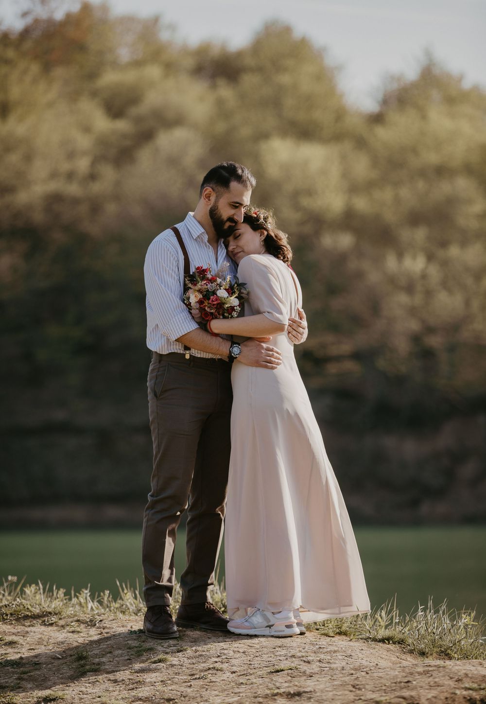 A couple holds each other close while standing in a meadow with a floral bouquet.