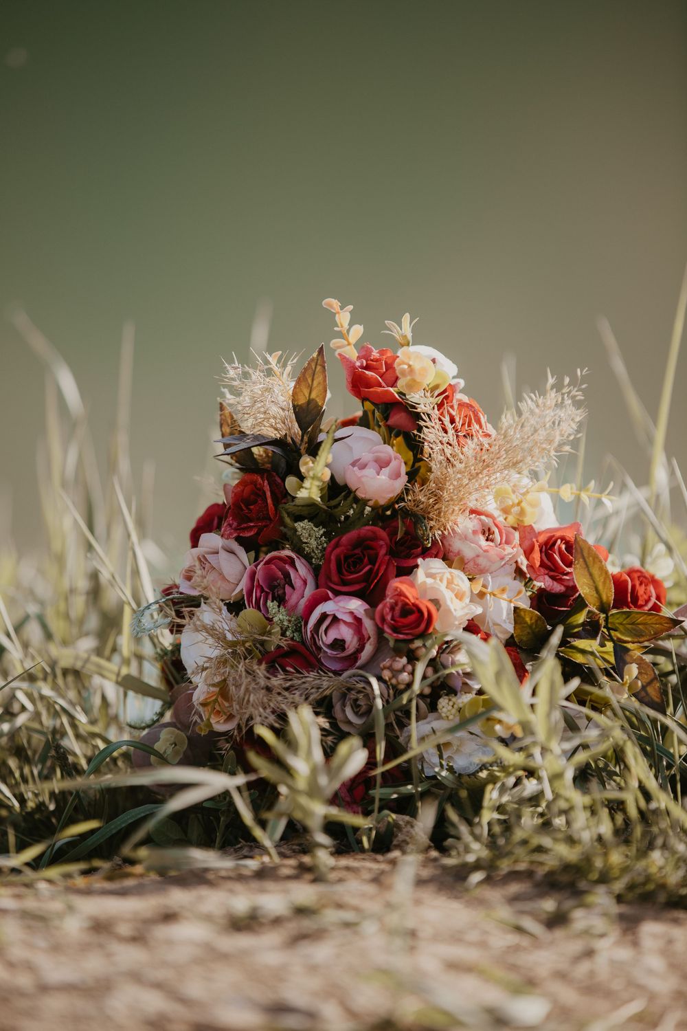 A lush bouquet of pink and red roses with dried flowers sits in tall grass.
