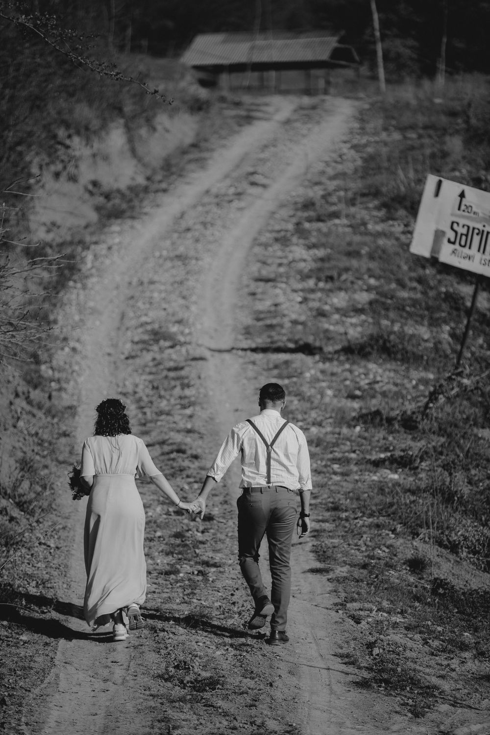 A couple walks hand in hand down a rural dirt path toward a distant building.