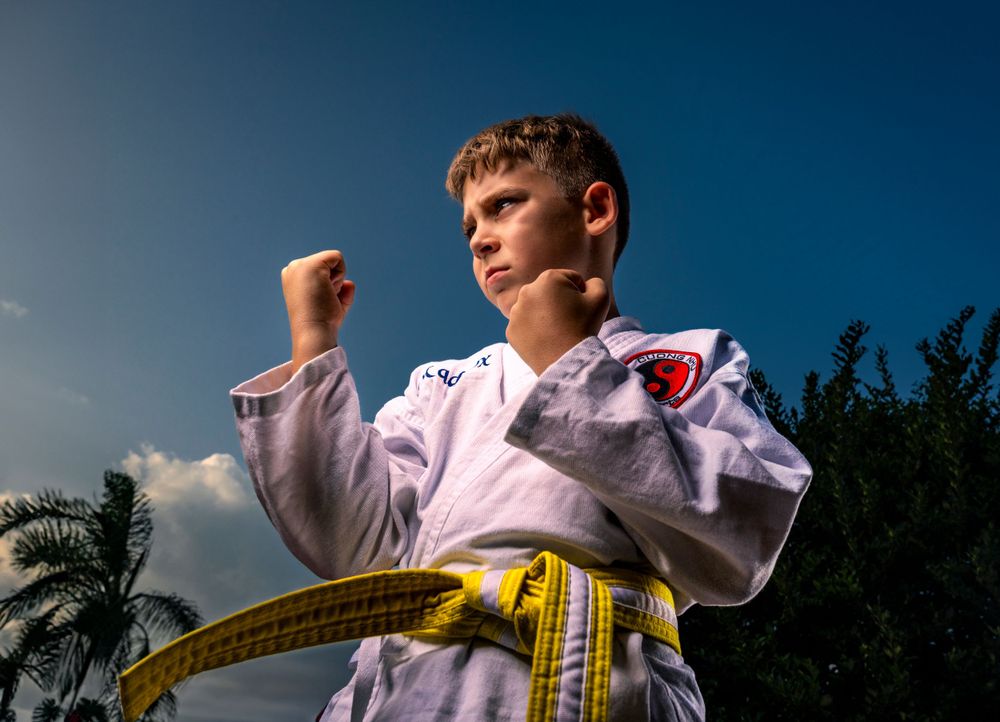 A young martial artist in a white karate gi with yellow belt strikes a powerful stance against a blue sky.