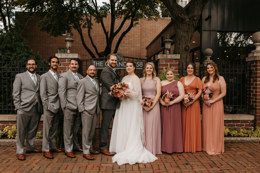 Wedding party poses in coordinated mauve and peach gowns with gray suits outdoors.