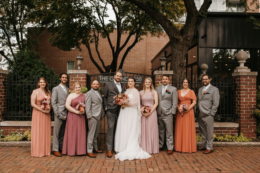 Bridal party in mismatched warm-toned dresses and gray suits pose before brick building.