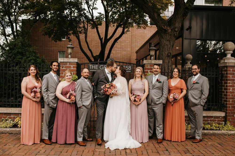 Bride and groom kiss surrounded by wedding party in coral dresses and gray suits.