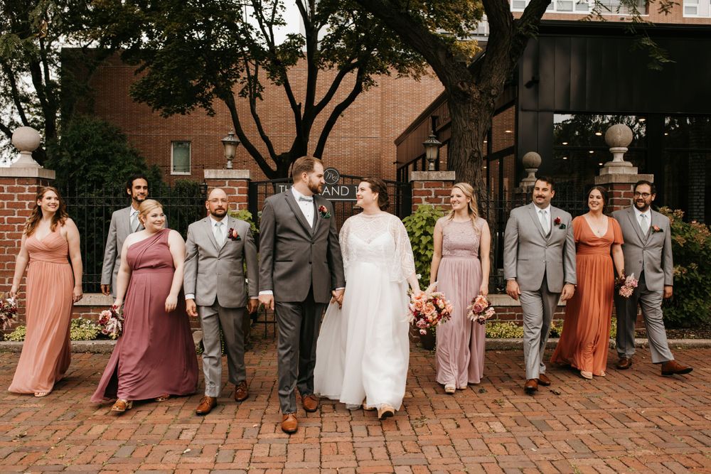 Wedding party walks together holding hands on brick pathway in warm autumn attire.