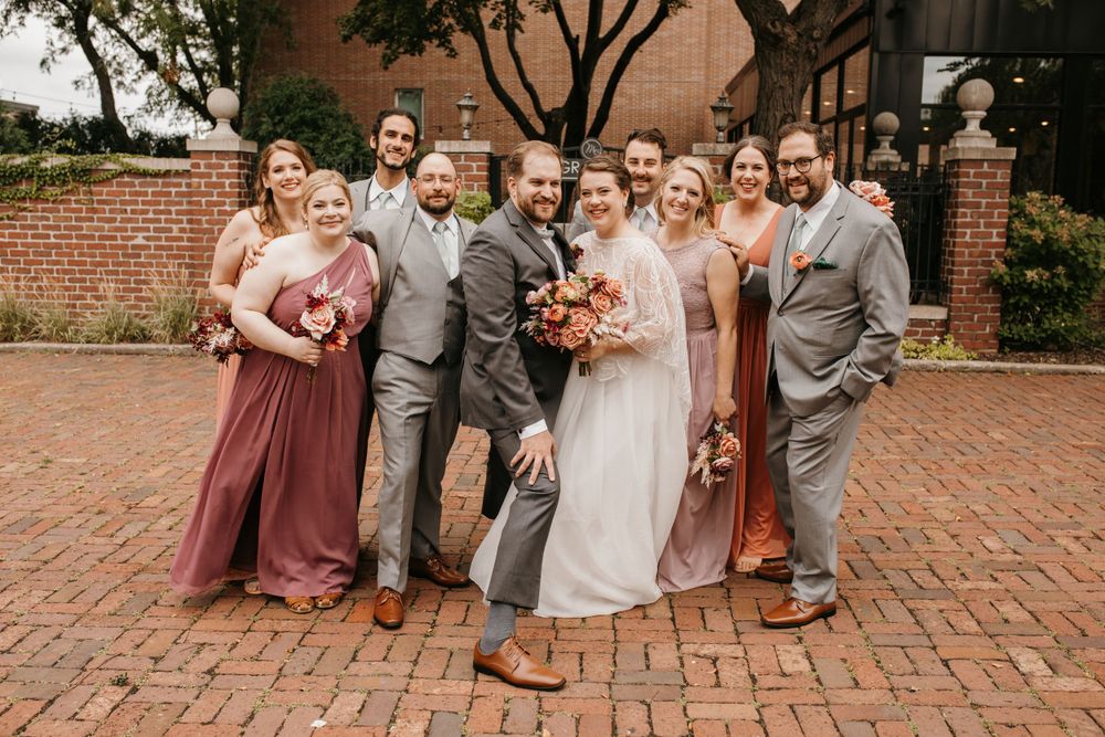 Joyful wedding party laughs and poses together casually on brick patio outdoors.