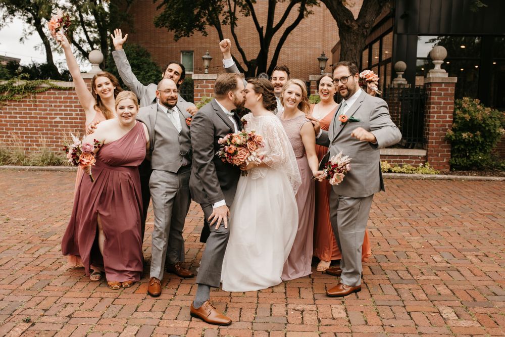 Wedding party celebrating on brick pathway, bridesmaids in rust and mauve gowns with groomsmen in gray suits kissing couple.