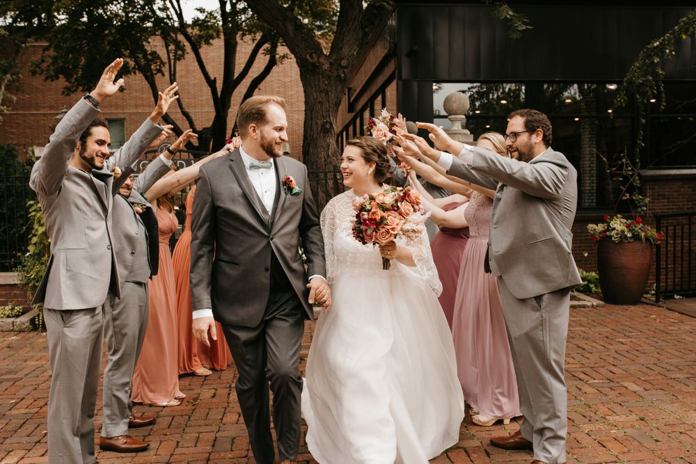 Bride and groom walk through tunnel of raised hands formed by wedding party in gray suits and earth-tone bridesmaid dresses.