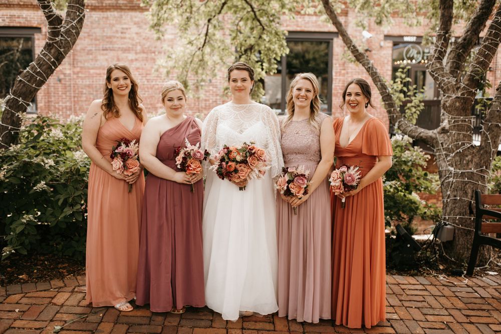 Bride in white lace gown poses with four bridesmaids in mismatched rust, mauve, and blush floor-length dresses outdoors.