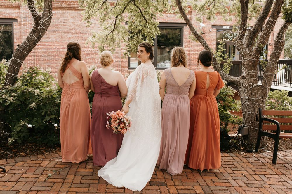 Bride and four bridesmaids in earth-tone gowns viewed from behind against a brick wall surrounded by blooming trees.