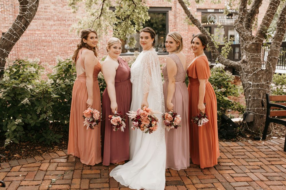 Bride in white cape gown poses with four bridesmaids in terracotta and mauve dresses holding bouquets in garden setting.