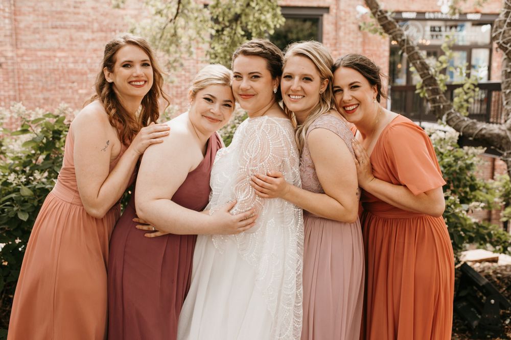 Bride in lace gown laughs with four bridesmaids in rust and blush dresses embracing in an outdoor garden setting.