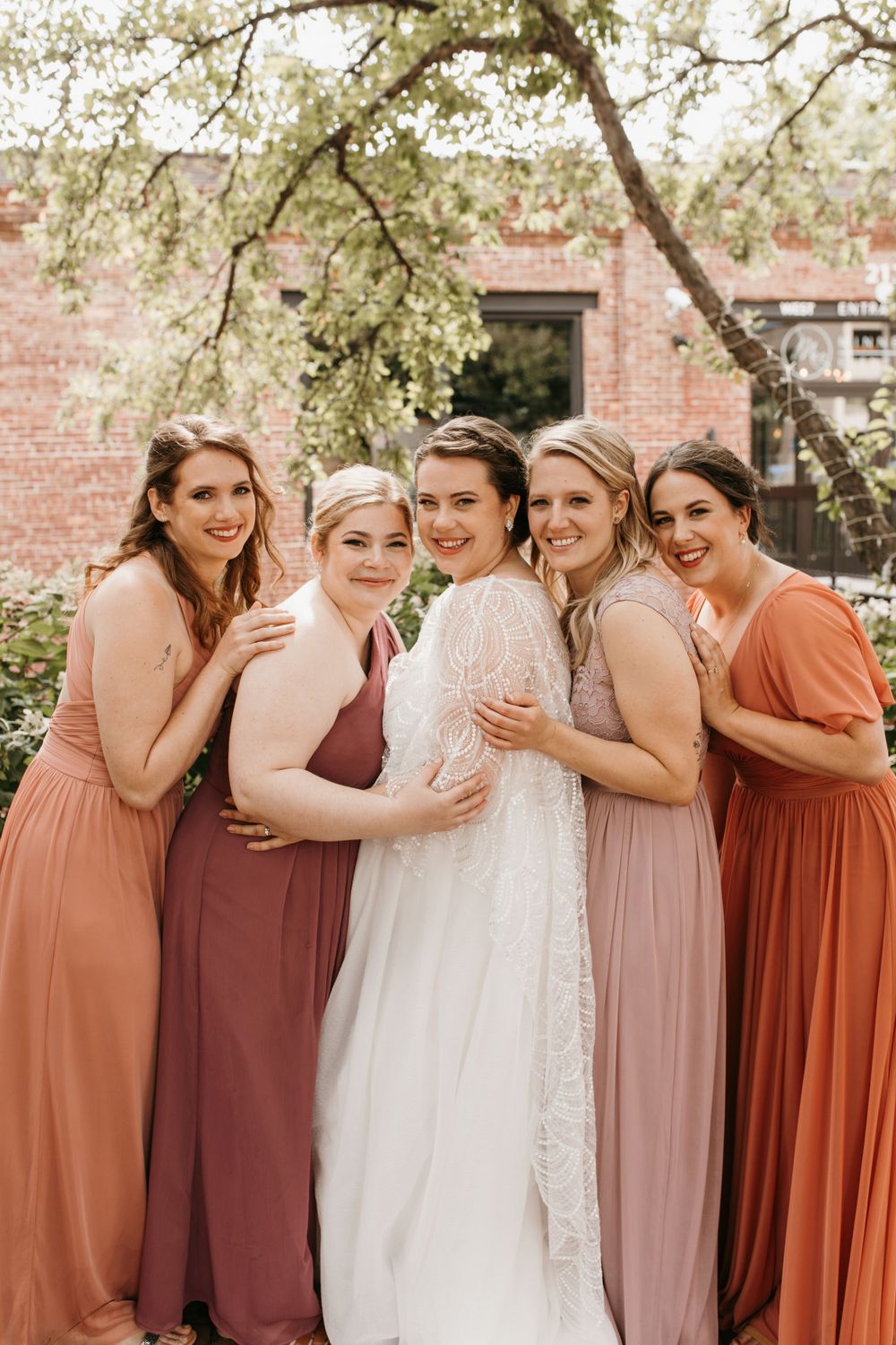 Five women smile together outdoors, bride in white lace gown surrounded by bridesmaids in rust and mauve floor-length dresses.