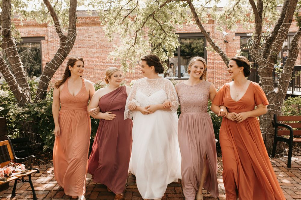 Bride and four bridesmaids in mismatched earth-tone gowns walk together laughing on brick pathway beneath flowering trees.