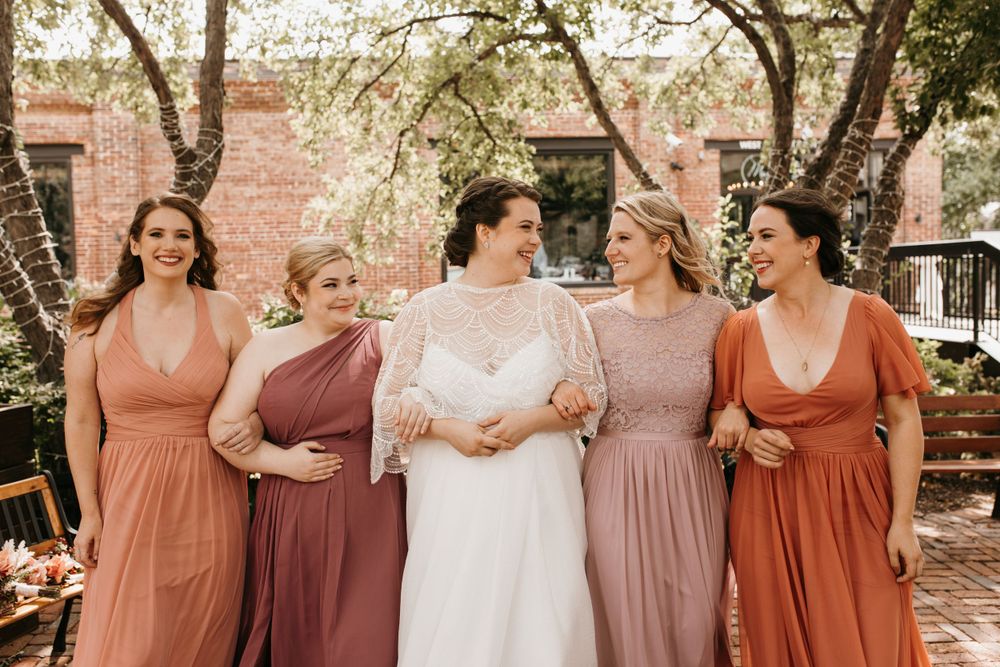 Bride in white lace gown laughs with four bridesmaids in terracotta and dusty rose dresses in outdoor garden setting.