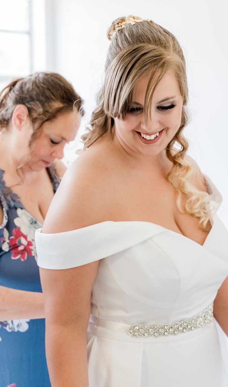 A bride in an off-shoulder white wedding gown with crystal belt detail smiles during preparations.