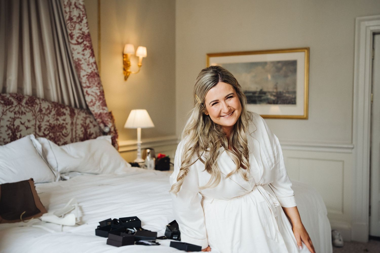 Bride in white robe sitting on luxurious hotel bed during wedding preparations.