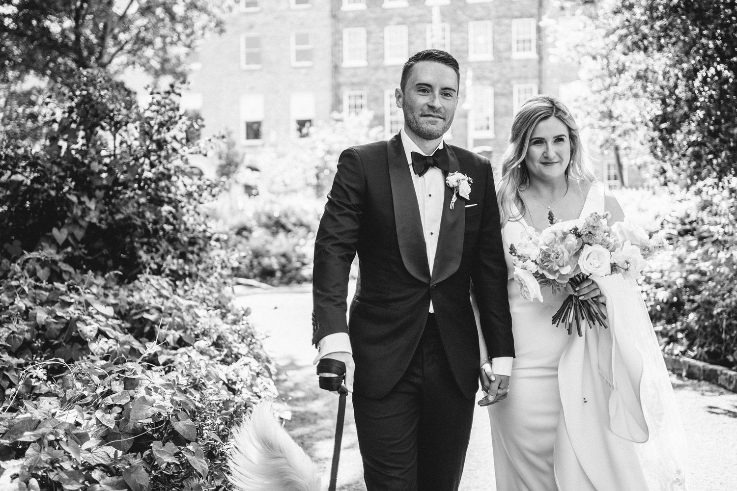 A black and white wedding photo of a couple walking together in a garden setting.
