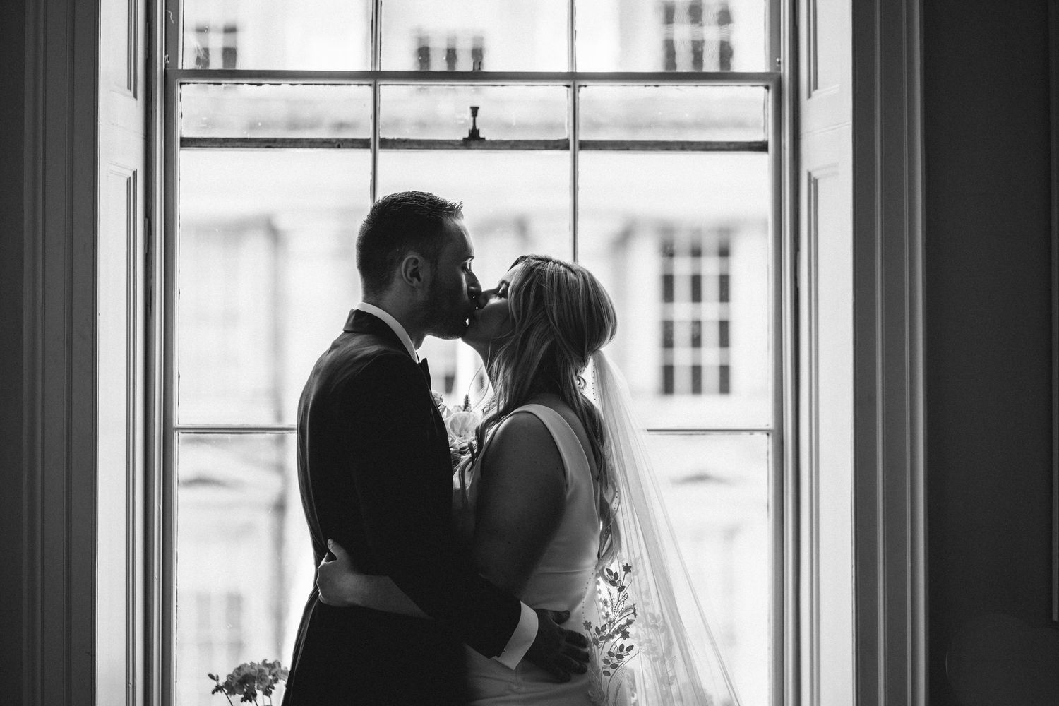 A silhouetted black and white wedding portrait of a couple kissing by a window.