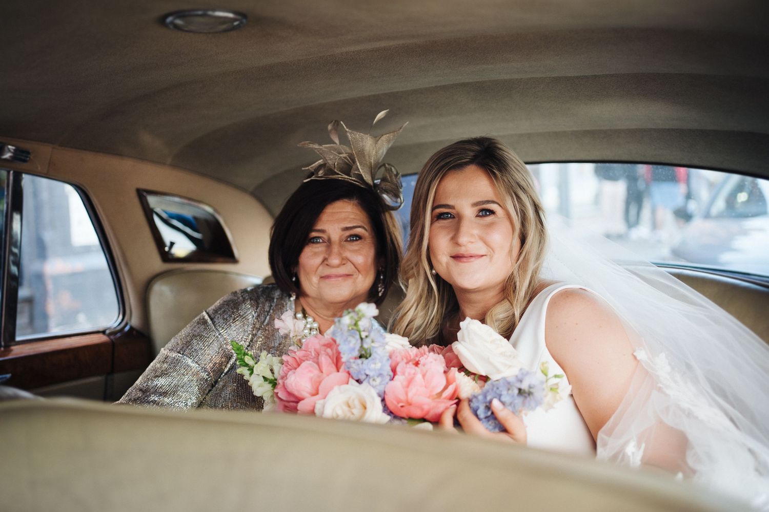 Two wedding attendees share a moment in the back seat of a vintage car with floral bouquets.