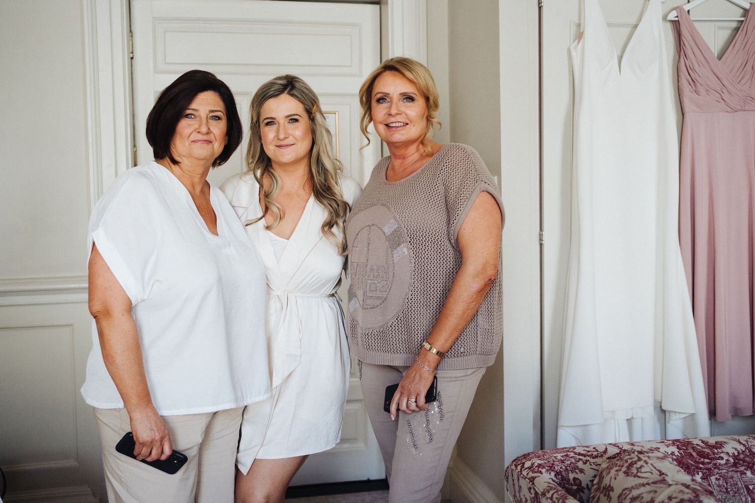 Three women in white and gray outfits pose together in an elegant room with a pink dress hanging in the background.