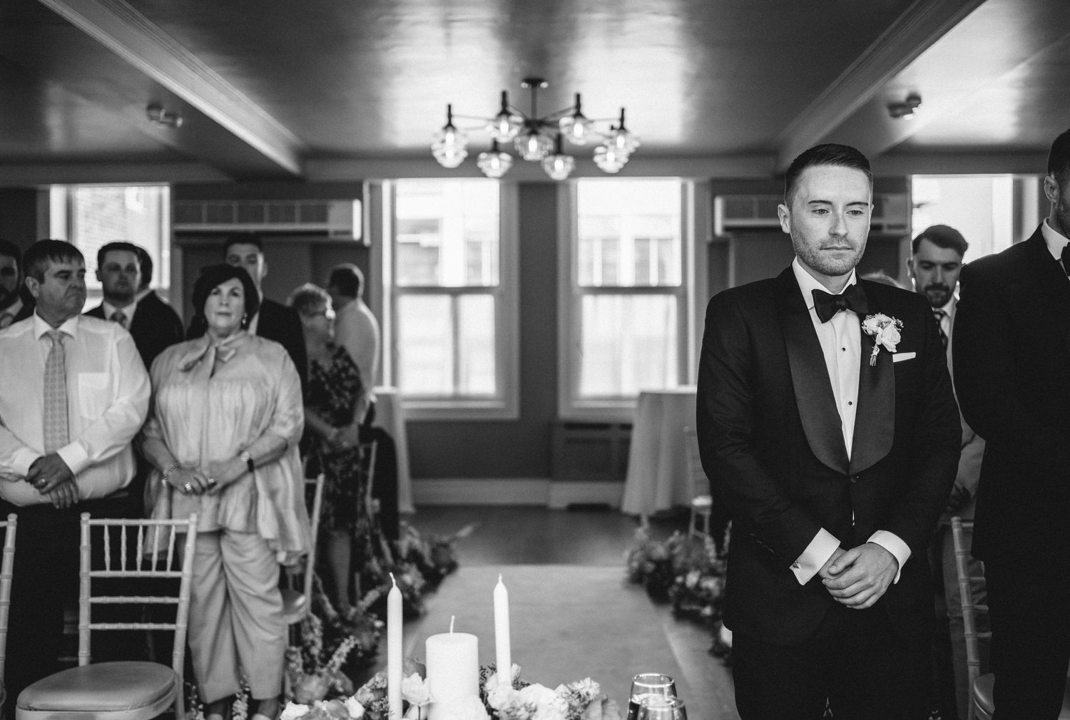 A black and white wedding ceremony photo showing guests and a groom waiting in an elegant room with chandeliers.